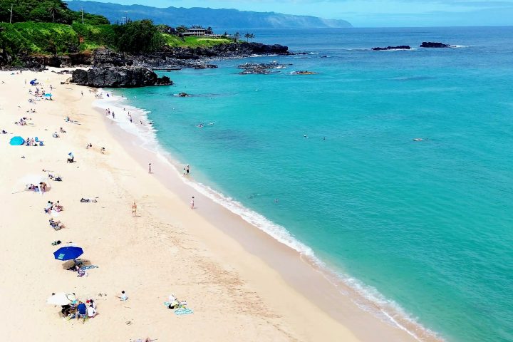 Waimea Bay’s sandy shoreline and turquoise water on a calm day, with rocky coastline beyond